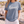 A woman standing on a sandy beach wearing a slate blue t-shirt with a white graphic print that reads 'Redeemed' and 'FAITH AS IS'.