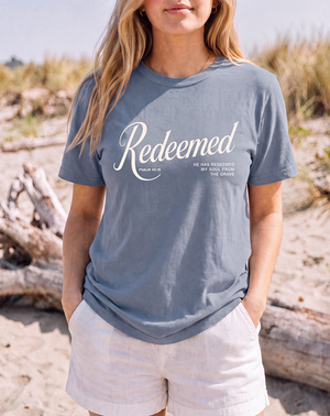 A woman standing on a sandy beach wearing a slate blue t-shirt with a white graphic print that reads 'Redeemed' and 'FAITH AS IS'.