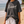 A woman standing on a sandy beach wearing a faded dark charcoal gray graphic t-shirt with 'Redeemed' printed in white script, paired with white shorts and holding a black Bible.