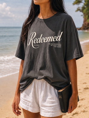 A woman standing on a sandy beach wearing a faded dark charcoal gray graphic t-shirt with 'Redeemed' printed in white script, paired with white shorts and holding a black Bible.