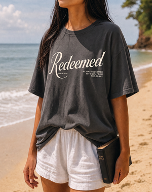A woman standing on a sandy beach wearing a faded dark charcoal gray graphic t-shirt with 'Redeemed' printed in white script, paired with white shorts and holding a black Bible.